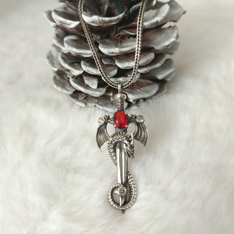May include: A silver pendant necklace featuring a dragon wrapped around a sword with a red gemstone. The pendant is hanging from a silver chain.