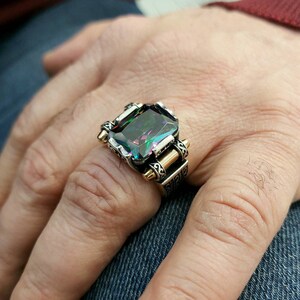 May include: A silver ring with a large, emerald-cut gemstone. The gemstone has a dark green base with iridescent rainbow colors. The ring has a detailed, ornate design with gold accents.