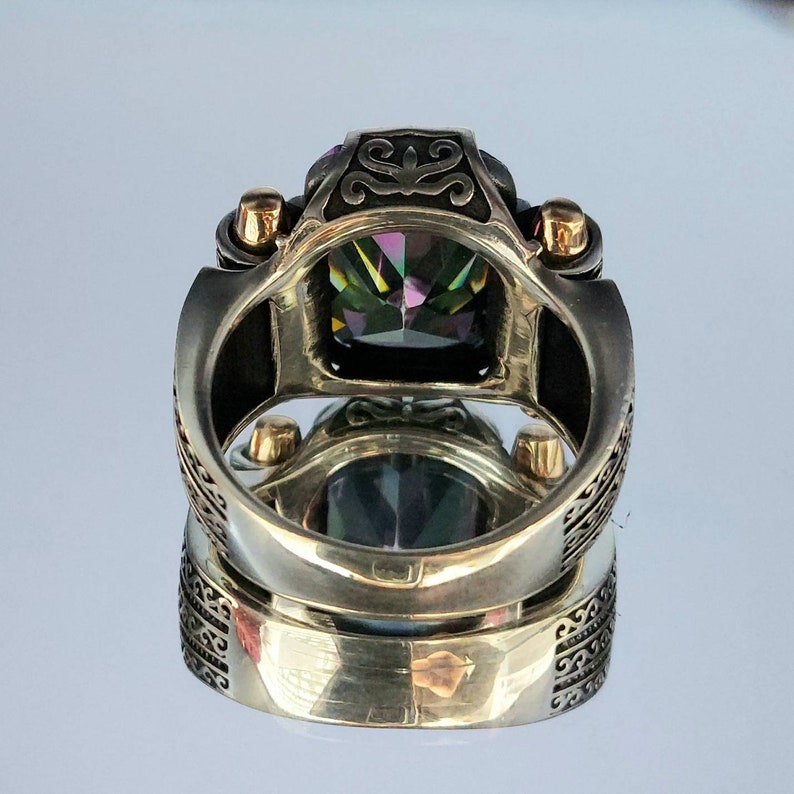 May include: A silver ring with a large, faceted, iridescent gemstone set in a square bezel. The ring has a wide band with intricate engraved details and gold accents.