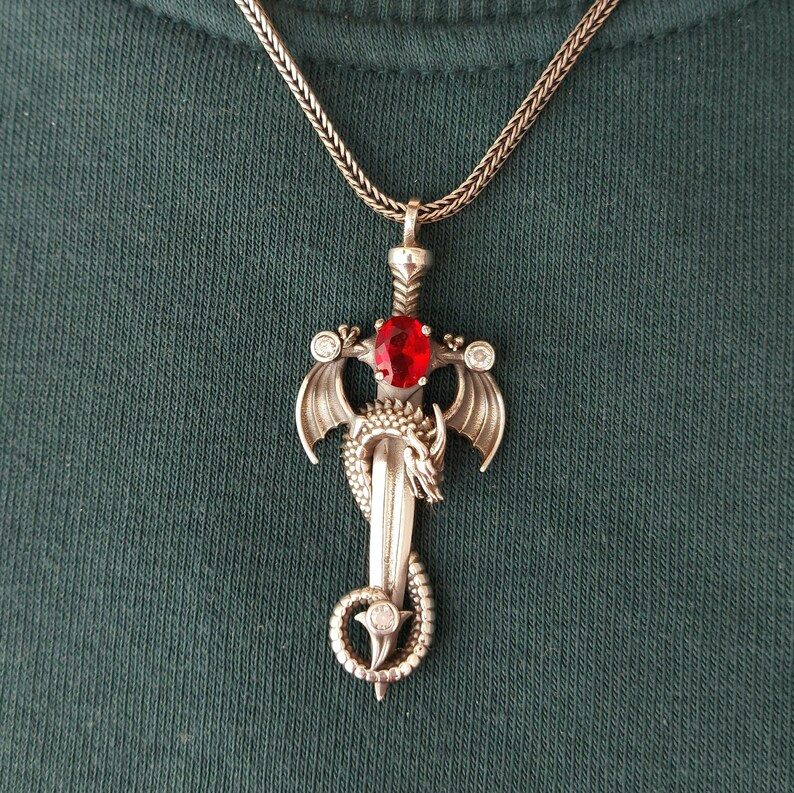 May include: A silver dragon sword pendant necklace with a red gemstone. The pendant features a detailed dragon design with wings and a sword.