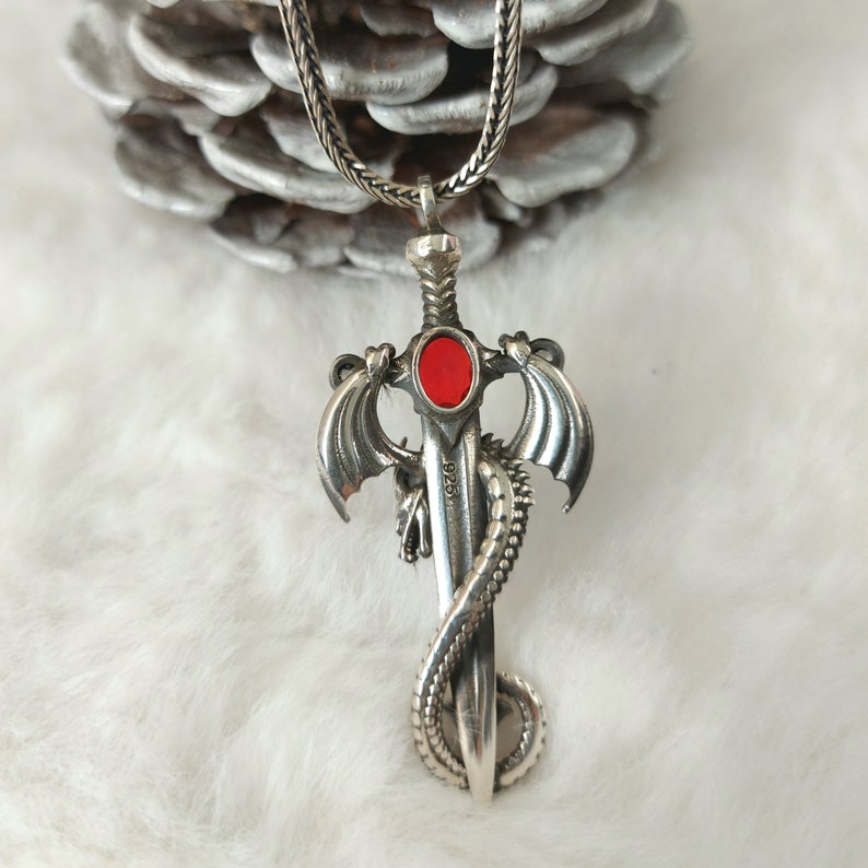 May include: A silver pendant necklace featuring a dragon wrapped around a sword with a red gemstone. The pendant has the inscription "925" on the sword.