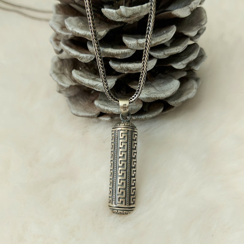 May include: A silver chain necklace with a silver pendant. The pendant is a cylinder with a geometric pattern. The pendant is marked with the number "925".
