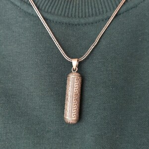 May include: A silver chain necklace with a silver pendant. The pendant is cylindrical and has a geometric pattern.
