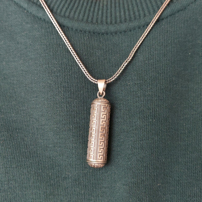 May include: A silver chain necklace with a silver pendant. The pendant is cylindrical and has a geometric pattern.