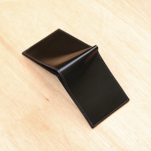 May include: A black leather wallet with a glossy finish, open to reveal the interior. The wallet is rectangular and has visible stitching around the edges. It rests on a light-coloured wooden surface.