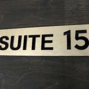 Puede incluir: Un letrero de metal dorado cepillado con letras negras que dice "SUITE 15".