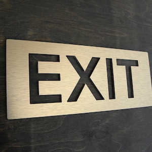 Op de afbeelding: Een rechthoekig bord in messingkleur met het woord "EXIT" uitgesneden. Het bord is gemonteerd op een donkere houten ondergrond. De letters zijn vetgedrukt en gemakkelijk te lezen, waardoor het een duidelijk richtingsbord is.