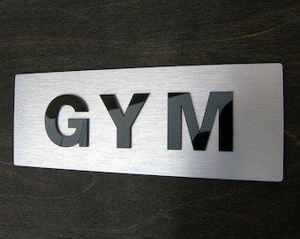 Op de afbeelding: Een geborsteld zilveren metalen bord met het woord "GYM" in zwarte reliëfletters.