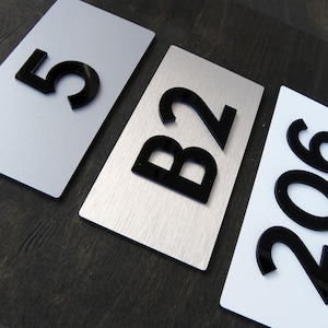 Puede incluir: Tres placas de dirección rectangulares con números y letras negros. Una placa es plateada con el número 5, otra es plateada con las letras B2, y la tercera es blanca con los números 206.