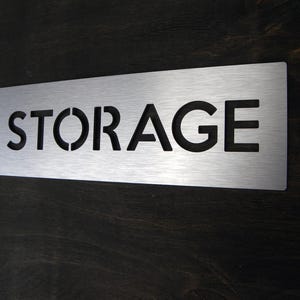 以下が含まれることがあります： 黒い文字で「STORAGE」とカットされた銀色の金属製の看板です。