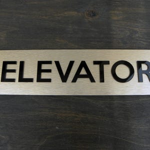 Może przedstawiać: Prostokątny znak ze szczotkowanego srebra z czarnymi wypukłymi literami, na którym widnieje napis "ELEVATOR".