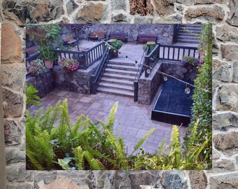 May include: A scenic outdoor courtyard with stone walls and stairs, framed by lush green ferns. The image is a wall mural, with a yellow chair, a wooden chair, and a white chair in the foreground, along with a wooden table with glass pitchers and potted plants.