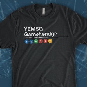 May include: Dark gray t-shirt with the text "YEMSG Gamehendge" in white. Below the text are six colored circles: blue, teal, green, red, orange, and yellow. The shirt is a crew neck.