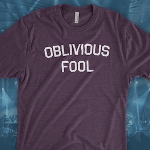 May include: A purple t-shirt with the words "OBLIVIOUS FOOL" printed in white block letters. The shirt has a crew neck and short sleeves. The background is a blurred image of a stage.