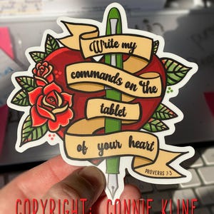 May include: A red heart with a green pen piercing through it. The pen has a gold nib and is wrapped in a yellow banner with the text "Write my commands on the tablet of your heart" and "Proverbs 7:3".