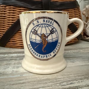 May include: Cream-colored ceramic mug with a gold rim and a handle. The mug features a circular emblem with the text "U.S. NAVAL OCEANOGRAPHIC OFFICE" and a seahorse illustration. The mug is set against a rustic wooden surface.