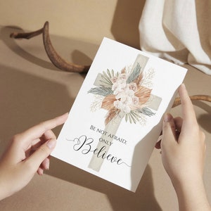 May include: A white card with a beige cross decorated with dried flowers and greenery. The text on the card reads "Be not afraid; only believe."