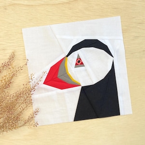 Puede incluir: Un cuadrado de tela blanco con un diseño geométrico negro, rojo, amarillo y gris que representa la cabeza de un frailecillo.