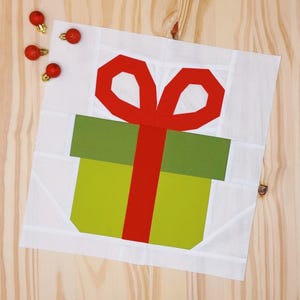 Könnte beinhalten: Ein weißer Stoffquadrat mit einer grünen und gelben Geschenkbox mit einer roten Schleife oben drauf.