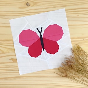 Puede incluir: Un cuadrado de tela blanco con un diseño de mariposa rosa y roja. La mariposa tiene un cuerpo negro y un contorno blanco.