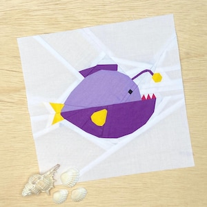 Puede incluir: Un pez pescador de papel morado y amarillo con fondo blanco. El pez tiene un señuelo amarillo y una boca roja.