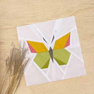 Pode incluir: Um quadrado de tecido branco com um design de borboleta geométrico. A borboleta é feita de peças de tecido amarelo, verde, rosa e cinza.