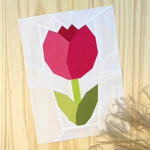 May include: A pink and green paper pieced tulip flower block. The tulip is made of pink and green fabric squares sewn together to create a geometric design.