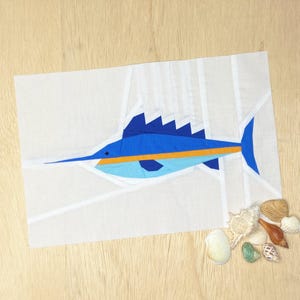 May include: A white fabric art piece featuring a blue marlin with an orange stripe. The marlin has a long, pointed snout and a distinctive dorsal fin. Various seashells are arranged on a wooden surface near the artwork.