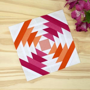 May include: A white fabric square with a pink and orange geometric pattern. The pattern is made up of triangles and rectangles arranged in a spiral design.