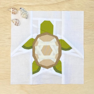 May include: A white fabric square with a green and brown paper turtle applique. The turtle has a geometric design with white lines.