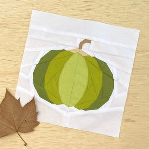 May include: A geometric paper piecing of a green pumpkin with a brown stem on a white background. A brown leaf rests on the wooden surface. The pumpkin is made of various shades of green paper.