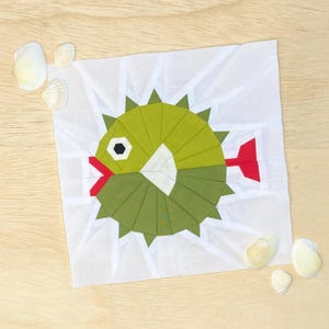 Puede incluir: Un bloque de colcha de pez globo verde y blanco con una aleta roja. El bloque está hecho de cuadrados de tela cosidos juntos en un patrón circular. El fondo es blanco.
