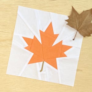 Pode incluir: Um quadrado de tecido branco com um desenho de folha de bordo de papel laranja no centro. A folha é feita de vários triângulos de papel.