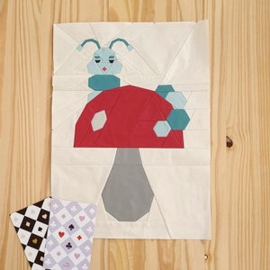 Pode incluir: Um painel de tecido branco com um cogumelo vermelho e cinza e uma lagarta azul e branca no topo. O cogumelo tem bolinhas brancas no topo. A lagarta tem um corpo azul e olhos brancos.