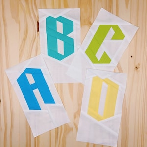 Puede incluir: Cuatro cuadrados de tela blanca con diferentes letras de colores. Las letras son A, B, C y D. Las letras están en un patrón geométrico y son en tonos de azul, verde y amarillo.