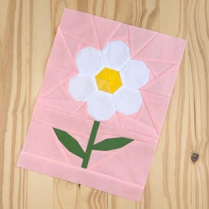 May include: A pink fabric square with a white flower and green stem. The flower has a yellow centre.