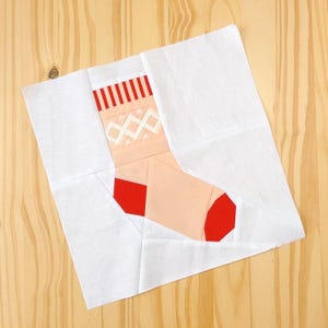 May include: A white fabric square with a red and white striped sock appliqué. The sock is pink with a white criss-cross pattern and red accents.