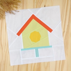 May include: A fabric quilt block with a house design. The house has a yellow body, an orange roof, and a yellow sun. The block is set against a white background with a light blue accent and is on a wooden surface.