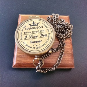 May include: A brass compass with a black inscription that reads "I hope you believe in yourself as much as I believe in you. GRANDSON Never forget that I Love You Forever". The compass is on a wooden base and has a chain attached.