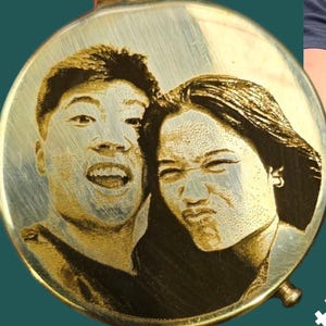 May include: A circular, gold-toned pendant featuring a laser-engraved image of two individuals. The faces display expressions of happiness and a humorous grimace. The pendant has a brushed metal finish and a small loop for a chain.
