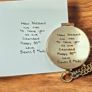 May include: A gold-toned, circular locket with a hinged lid and chain, beside a white card. Both the locket and card display handwritten text: "How blessed we are to have you as our Grandad Happy 80th Love, Beans & Mads."