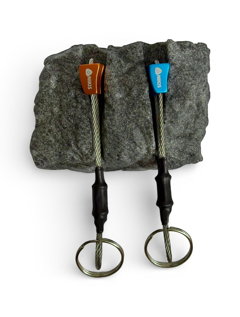 May include: Two climbing nuts with metal heads in orange and blue, each featuring the brand name NIKIS printed in white. The heads are attached to flexible steel cables with black heat-shrink tubing and circular metal rings at the ends.