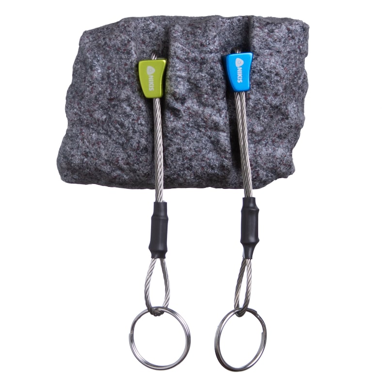May include: Two climbing nuts with wire cables and metal rings attached to a textured grey rock-like block. One nut is lime green and the other is blue, both featuring the text NIKIS printed in white. The cables are secured with black sleeves.
