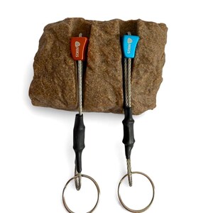 May include: Two climbing nuts, one red and one blue, each labelled with the text NIKIS. The metal nuts are attached to steel cables with black heat-shrink tubing and silver circular rings, positioned within a textured brown rock-like block.