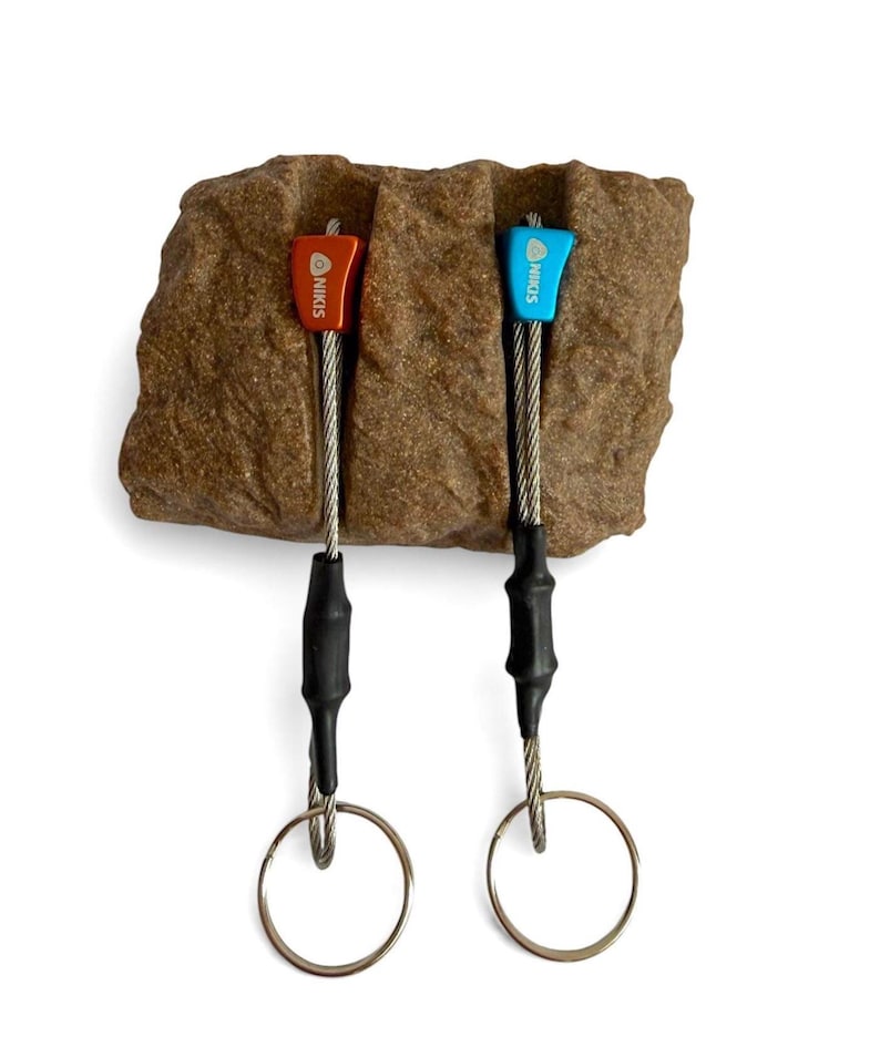 May include: Two climbing nuts, one red and one blue, each labelled with the text NIKIS. The metal nuts are attached to steel cables with black heat-shrink tubing and silver circular rings, positioned within a textured brown rock-like block.