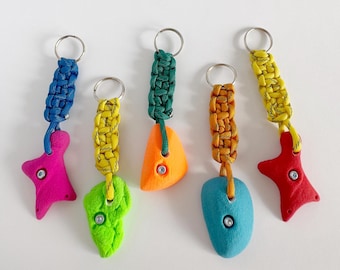 May include: Five colorful climbing hold keychains. Each keychain features a different colored climbing hold, including pink, green, orange, blue, and red. The holds are attached to braided cords in various colors, with a silver keyring at the top.
