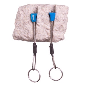 May include: Two blue metal climbing nuts with the brand name NIKIS printed on them, each attached to a flexible steel cable with a black heat-shrink sleeve and a circular metal ring at the end, all inserted into a textured beige rock block.