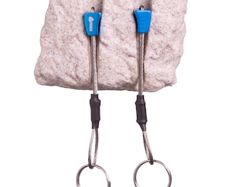 May include: Two blue metal climbing nuts with the brand name NIKIS printed on them, each attached to a flexible steel cable with a black heat-shrink sleeve and a circular metal ring at the end, all inserted into a textured beige rock block.