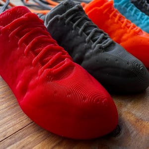 Może przedstawiać: Rząd kolorowych, drukowanych w 3D miniaturowych sneakersów w kolorach czerwonym, szarym, pomarańczowym, turkusowym, czarnym i pomarańczowym. Buty ułożone są na drewnianej powierzchni, z widocznym brelokiem i sznurkami. Unikalny przedmiot dekoracyjny.