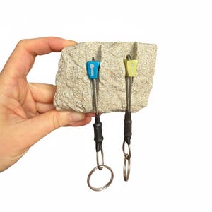 May include: A light blue and a light green climbing nut, each attached to a steel cable with a black heat-shrink sleeve and a metal ring, inserted into vertical grooves on a rectangular piece of textured, light beige granite rock.